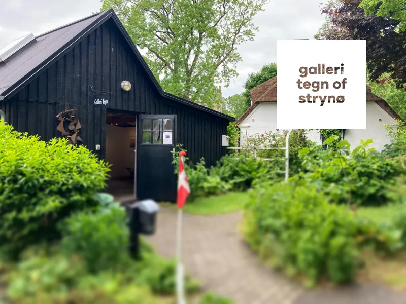 Den sorte træbygning med Galleri Tegns hvide skilt, en dansk vimpel og en grøn have foran