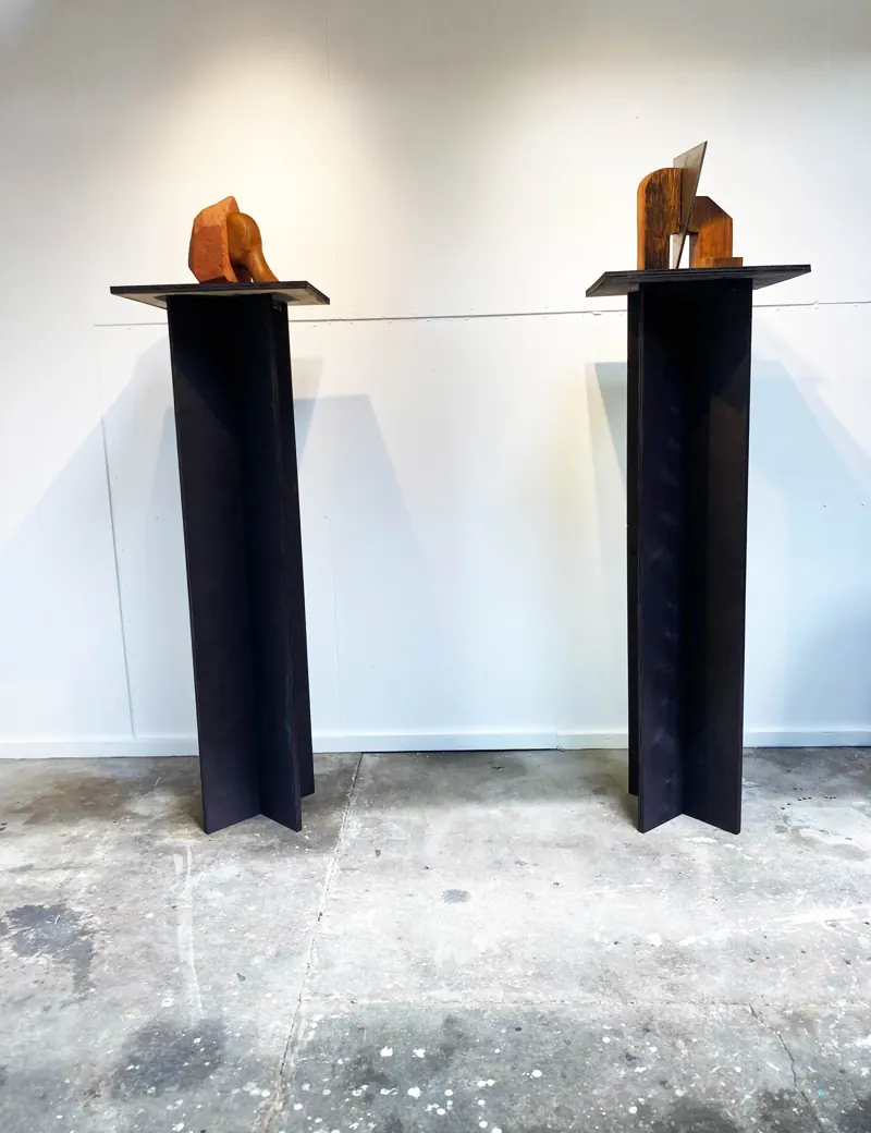 Two black steel plinths with wooden forms, seen against a white wall