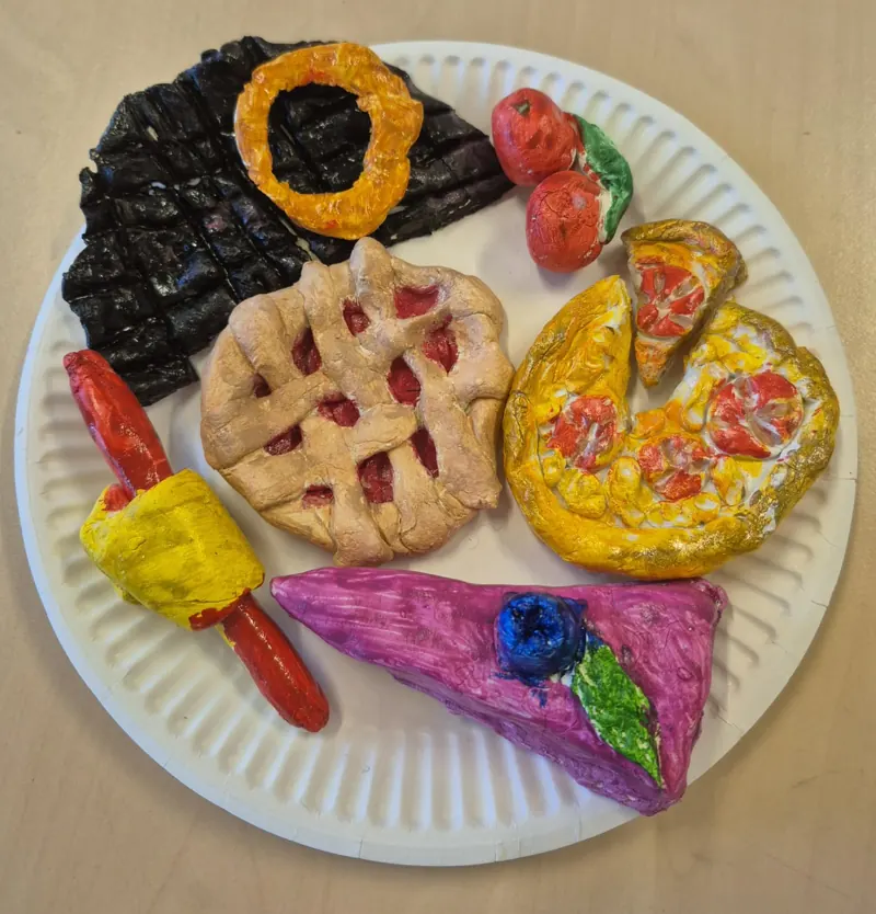Painted clay food on a paper plate: a pie, a pizza slice, a sausage and a doughnut